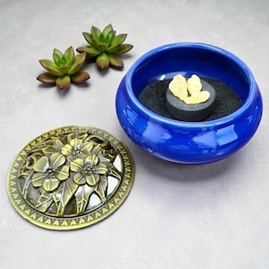 May include: A cobalt blue ceramic bowl containing a black charcoal disc topped with pale yellow resin pieces. Beside the bowl lies a circular, antique brass-colored metal lid featuring a raised floral relief pattern and two small green succulents.