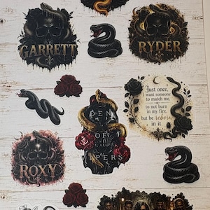 Può includere: Un foglio di adesivi con vari disegni a tema scuro tra cui teschi, serpenti, rose e una mazza da baseball. Il testo include nomi come DIESEL, KENZO, GARRETT, RYDER, ROXY e frasi come BORN FROM DARKNESS, CROWNED IN GOLD e VIPERS GIRL.