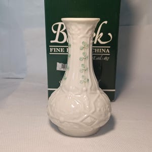May include: A white ceramic vase with a bulbous base and a slender neck. The vase is decorated with embossed patterns and green shamrock designs. The box behind the vase reads "Belleek Fine China Estd. 1857".
