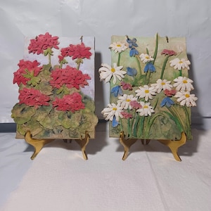 May include: Two decorative floral relief plaques. One features red geraniums, the other daisies, blue and pink flowers. Both are mounted on gold-colored stands. The plaques have a textured, painted appearance.