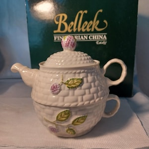 May include: A white porcelain teacup and teapot set with a basket weave design. The teapot has a lid with a pink berry on top and is decorated with green leaves and pink berries. The set is from Belleek Fine Parian China.