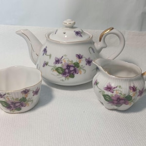 May include: A white porcelain tea set featuring a teapot, creamer, and sugar bowl. The set is decorated with purple violets and green leaves. The teapot has a gold handle and trim. The sugar bowl has a scalloped edge.