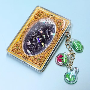 Puede incluir: Un llavero rectangular transparente con borde dorado y un óvalo central lleno de purpurina. Adjuntos hay colgantes con forma de gema verde, manzana roja y botella de poción verde, todos conectados por una cadena dorada.