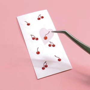 Puede incluir: Una hoja de pegatinas de cerezas con borde en forma de corazón. Las pegatinas son rojas con tallos verdes. Unas pinzas sujetan una pegatina. El fondo es de color rosa liso.