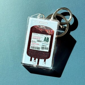 Puede incluir: Un llavero acr&iacute;lico transparente con un dise&ntilde;o de bolsa de sangre, con la inscripci&oacute;n "AB Positive". La bolsa est&aacute; llena de l&iacute;quido rojo oscuro e incluye texto y un c&oacute;digo de barras. Llavero con un aro met&aacute;lico plateado.