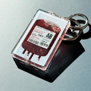 Puede incluir: Un llavero acr&iacute;lico transparente con un dise&ntilde;o de bolsa de sangre con purpurina roja. El llavero tiene un llavero plateado y una etiqueta impresa con el texto "AB Rh Positive". El dise&ntilde;o imita una bolsa de sangre.