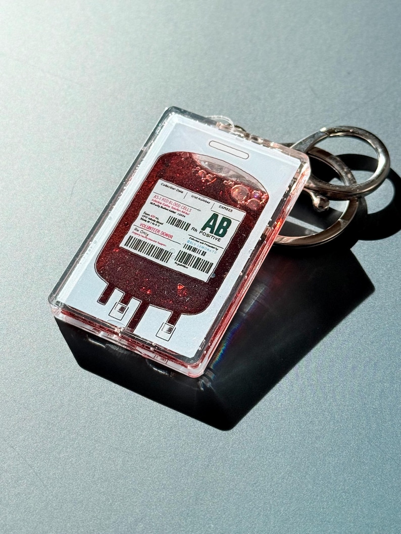 Puede incluir: Un llavero acr&iacute;lico transparente con un dise&ntilde;o de bolsa de sangre con purpurina roja. El llavero tiene un llavero plateado y una etiqueta impresa con el texto "AB Rh Positive". El dise&ntilde;o imita una bolsa de sangre.