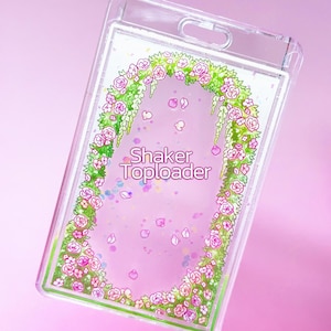 May include: Clear plastic Shaker Toploader with a floral design. The toploader features a wreath of pink roses and green leaves, with floating pink petals and the words "Shaker Toploader". The background is a soft pink.