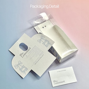 Puede incluir: Detalle del embalaje de un producto. La caja es blanca con una tapa transparente. El embalaje incluye una tarjeta blanca plegada con un orificio para el asa y una peque&ntilde;a tarjeta blanca con la marca "blu. Lemonayd". El fondo es un degradado de rosa y azul.