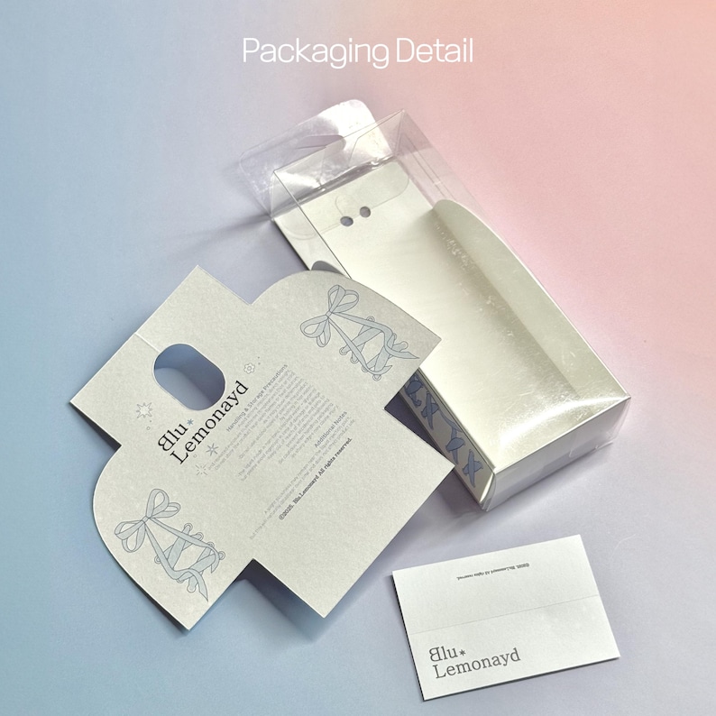 Puede incluir: Detalle del embalaje de un producto. La caja es blanca con una tapa transparente. El embalaje incluye una tarjeta blanca plegada con un orificio para el asa y una peque&ntilde;a tarjeta blanca con la marca "blu. Lemonayd". El fondo es un degradado de rosa y azul.