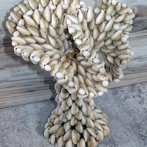 May include: A decorative sculpture made of numerous small, cream-colored seashells. The sculpture has a rounded top and a flared base, with a tag attached. The seashells are tightly packed together, creating a textured surface. The sculpture is on a gray surface.