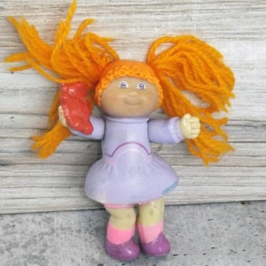 May include: A vintage doll with orange yarn hair styled in pigtails. The doll wears a lavender dress, pink socks, and purple shoes. It holds a red object in its left hand. The doll has a smiling face and blue eyes.