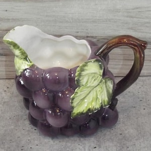 May include: A ceramic creamer pitcher shaped like a cluster of purple grapes with green leaves and a brown handle. The interior of the pitcher is white, and the exterior features a glossy finish with a textured surface resembling individual grapes.