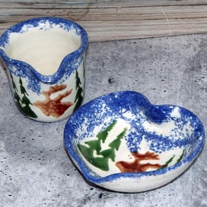 Puede incluir: Una cremaera de cerámica y un plato en forma de corazón. Ambos tienen un borde azul moteado y un interior blanco. La cremaera y el plato presentan una escena de bosque con árboles verdes y un ciervo marrón.