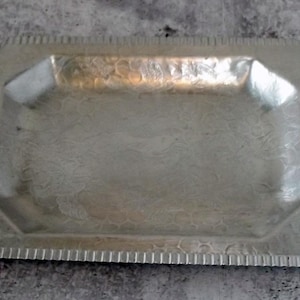May include: A rectangular silver-colored tray with a textured surface and a decorative floral pattern. The tray has a raised, octagonal central area and a scalloped edge. The tray is made of metal and is likely vintage.