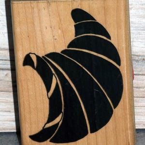 May include: A wooden stamp with a black croissant design. The stamp is rectangular and made of light-colored wood. The croissant is depicted in a stylized, graphic manner, with curved lines and a solid black fill.