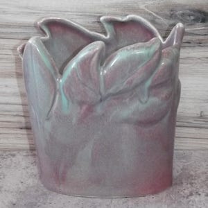 Puede incluir: Un jarrón de cerámica con un diseño único en forma de pétalos en el borde. El jarrón presenta una mezcla de colores rosa, gris y turquesa, creando un efecto marmolado. El jarrón es cilíndrico y se apoya sobre una base plana.