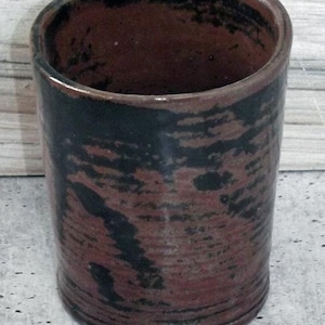 May include: A cylindrical ceramic cup with a dark brown interior and exterior. The exterior features a mottled pattern of black and reddish-brown hues. The cup has a slightly flared rim and a smooth, glossy finish. The cup is approximately 4 inches tall.