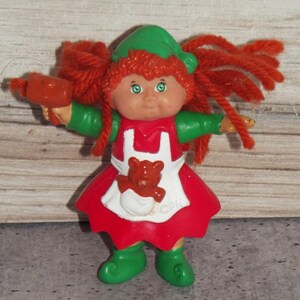 May include: A small Cabbage Patch Kids figurine with bright orange yarn hair, a green hat, and green shoes. The figure wears a red dress with a white apron featuring a brown teddy bear graphic and the letters CPK. It holds a small red object.