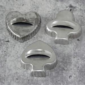 May include: Three vintage metal cookie cutters in the shapes of a heart, a club, and a spade. Each cutter has a handle and a scalloped edge. The cutters are made of a silver-colored metal and are arranged on a gray surface.