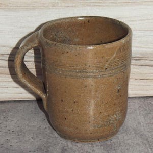 May include: A cylindrical ceramic mug with a speckled tan and brown glaze. The mug features a thick, curved handle on the left side and two horizontal incised lines encircling the upper portion of the body, creating a textured, earthy appearance.