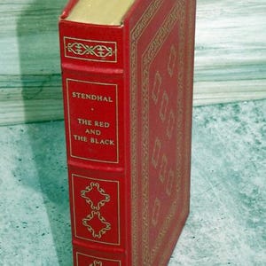May include: A red hardcover book titled "The Red and the Black" by Stendhal. The book has gold decorative detailing and gold page edges. The spine displays the author's name and title in gold lettering.
