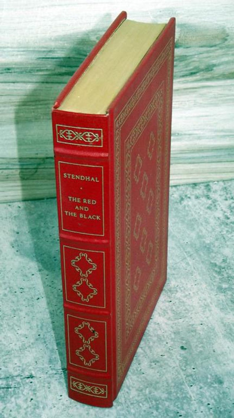 May include: A red hardcover book titled "The Red and the Black" by Stendhal. The book has gold decorative detailing and gold page edges. The spine displays the author's name and title in gold lettering.