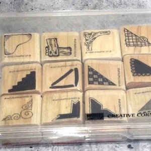 May include: A clear plastic storage container holding twelve square wooden rubber stamps. Each stamp features a unique black ink corner design, including scrolls, geometric patterns, and lines. The text "CREATIVE CORNERS" is printed on the case.