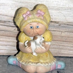 Puede incluir: Una figura de cerámica de un niño con cabello rubio peinado en dos coletas con lazos rosas, vistiendo un vestido amarillo con dobladillo con volantes. El niño tiene ojos azules y sostiene un pequeño cachorro blanco con manchas marrones.