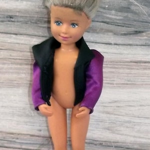 May include: A doll with grey hair and blue eyes wearing a black and purple jacket. The doll is standing on a light grey wooden surface. The doll's skin is a light peach colour.