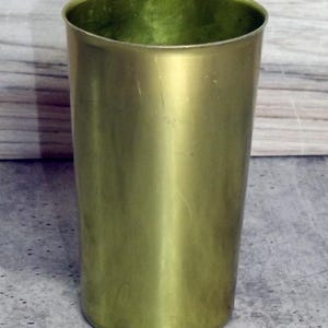 May include: A cylindrical drinking vessel with a smooth, metallic gold-toned finish. The cup features a simple, straight-sided design with a wide, open top and a flat base, showcasing a uniform, reflective surface throughout the exterior and interior.