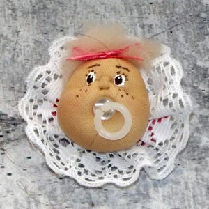 Puede incluir: Una cabeza de muñeca hecha a mano con un chupete. La muñeca tiene una cara bronceada con rasgos pintados, incluidas pecas, y un pequeño lazo rosa. Está rodeada por un cuello de encaje blanco.