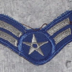 May include: A chevron-shaped embroidered patch featuring a blue background with two horizontal silver stripes and a central silver star enclosed within a circular border. The edges of the patch show visible loose threads and a textured fabric finish.