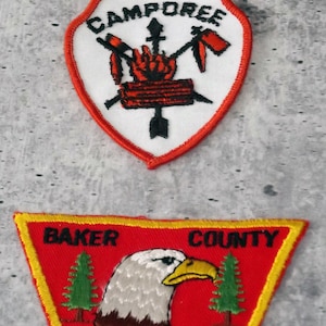 May include: Two embroidered patches. The top patch is shield-shaped with the word "CAMPOREE" and a design of crossed arrows, a fire, and a tomahawk. The bottom patch is rectangular with "BAKER COUNTY" and an eagle.