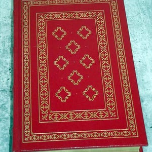 May include: A red hardback book with gold decorative detailing. The cover has a gold border and a central design of six diamond-shaped patterns. The book's spine is visible, and the pages have a gold edge.