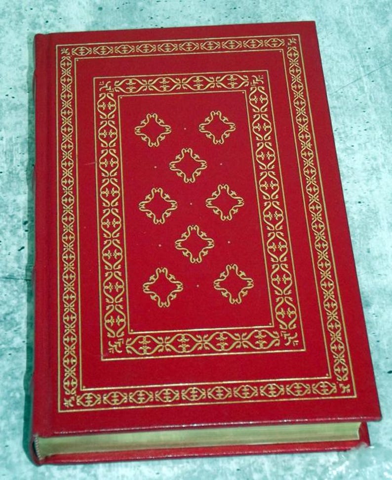 May include: A red hardback book with gold decorative detailing. The cover has a gold border and a central design of six diamond-shaped patterns. The book's spine is visible, and the pages have a gold edge.