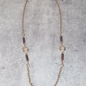 May include: A long gold-tone chain necklace featuring alternating dark brown rectangular links and circular gold-tone accents with a central clear stone. The necklace has a delicate link chain design that connects the decorative elements symmetrically.