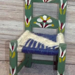 May include: A miniature green wooden chair with a woven seat of blue and white yarn. The chair is decorated with painted floral designs in white, red, and yellow. The chair's back and legs are painted green.