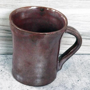May include: A handmade, maroon-colored ceramic mug with a curved handle. The mug has a glossy finish and a slightly irregular shape, suggesting it is handcrafted. The interior is dark, and the mug sits on a light-colored surface.