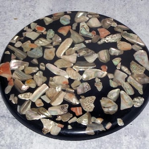 May include: A round, black decorative coaster inlaid with a mosaic of iridescent shell fragments in shades of white, beige, orange, and gray. The coaster has a glossy finish and a smooth, rounded edge. The shell pieces are irregularly shaped.