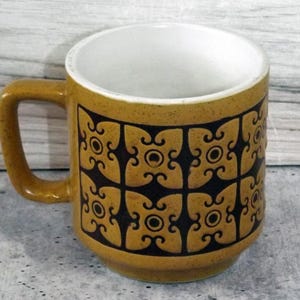 May include: Mustard yellow ceramic mug with a white interior. The mug features a repeating black geometric pattern. The handle is a matching mustard yellow. The mug is designed for hot beverages.