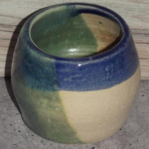 May include: A handmade ceramic bowl with a rounded shape. The bowl features a color-blocked design with sections of blue, green, and cream. The interior is a mix of green and brown. The bowl is approximately 4 inches in diameter and 3 inches tall.