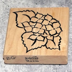 May include: A wooden stamp featuring a black floral design of hydrangea flowers and leaves. The stamp is made by Denami Design, with the company's logo and address visible. The stamp has the initials "BG" on the side.