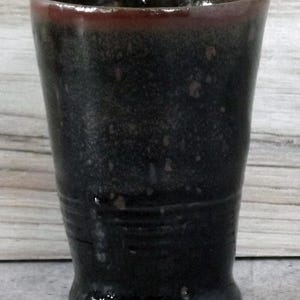 Puede incluir: Un vaso de cerámica hecho a mano con un esmalte oscuro y moteado. La taza tiene una forma acampanada con un borde marrón oscuro y una superficie ligeramente texturizada. La base es de un tono negro más oscuro. El vaso es adecuado para bebidas.