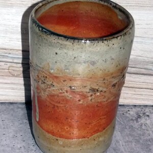 Puede incluir: Vaso de cerámica hecho a mano con un diseño texturizado único. La taza presenta una banda de esmalte naranja vibrante, que contrasta con los tonos beige y gris neutros. El interior es de color naranja oscuro.