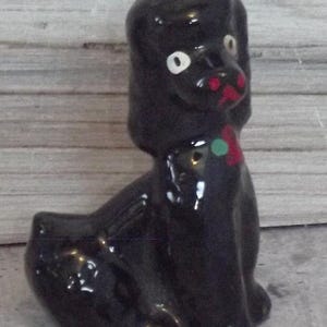 May include: A glossy black ceramic poodle figurine in a seated position. The dog features white eyes with black pupils, a red nose and mouth area, and a small green and red decorative accent on the chest area. The surface has a reflective finish.