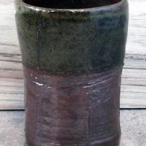 Puede incluir: Un vaso de cerámica hecho a mano con un esmalte de dos tonos. La parte superior es de un verde oliva oscuro, que pasa a un borgoña profundo en la base. La forma cilíndrica tiene sutiles líneas verticales, agregando textura.