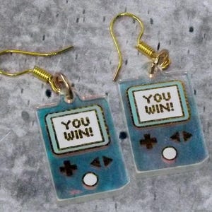 Puede incluir: Un par de pendientes originales con un diseño de consola de juegos portátil azul. Cada pendiente tiene un gancho dorado y un dije rectangular con las palabras "YOU WIN!" en un cuadro blanco. El diseño de la consola incluye botones direccionales y un círculo blanco.