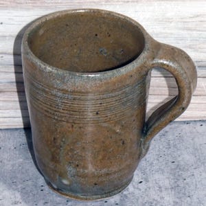 Puede incluir: Taza de cerámica marrón hecha a mano con asa curvada. La taza tiene una superficie texturizada con líneas horizontales en el centro. El interior es de un marrón más oscuro y el asa está integrada. Adecuada para bebidas frías o calientes.