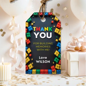 May include: A black gift tag with colorful building block accents and the words "THANK YOU" in yellow, green, and blue. The tag reads "FOR BUILDING MEMORIES WITH ME!" and "Love WILSON". The tag is tied with a brown string.